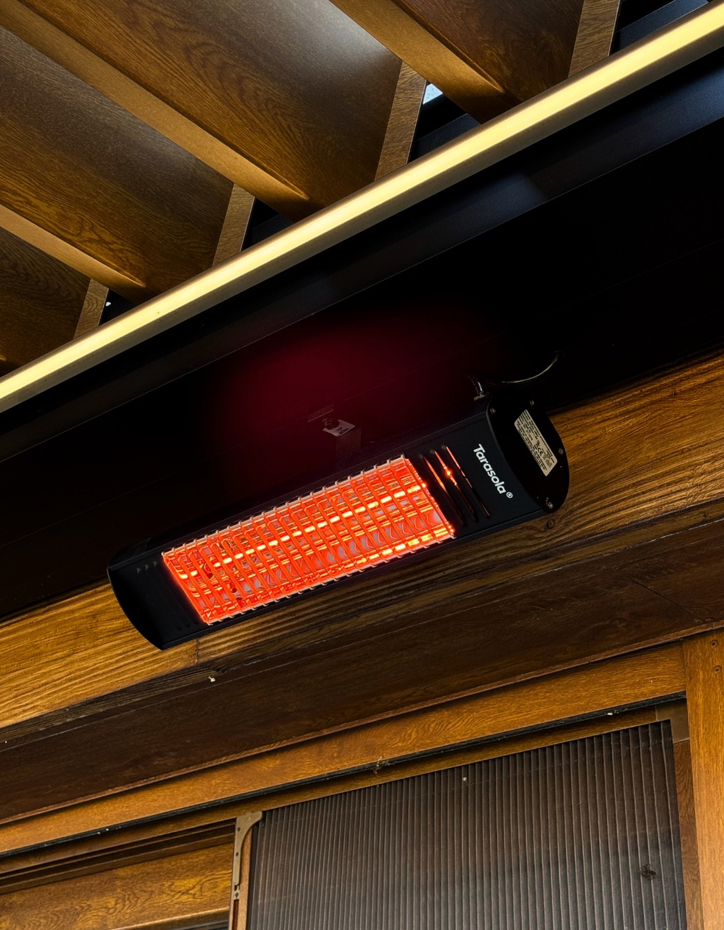 Czarny promiennik ciepła Tarasola zamontowany pod drewnianym sufitem z oświetleniem LED, widoczny fragment drewnianej ściany z żaluzjami.