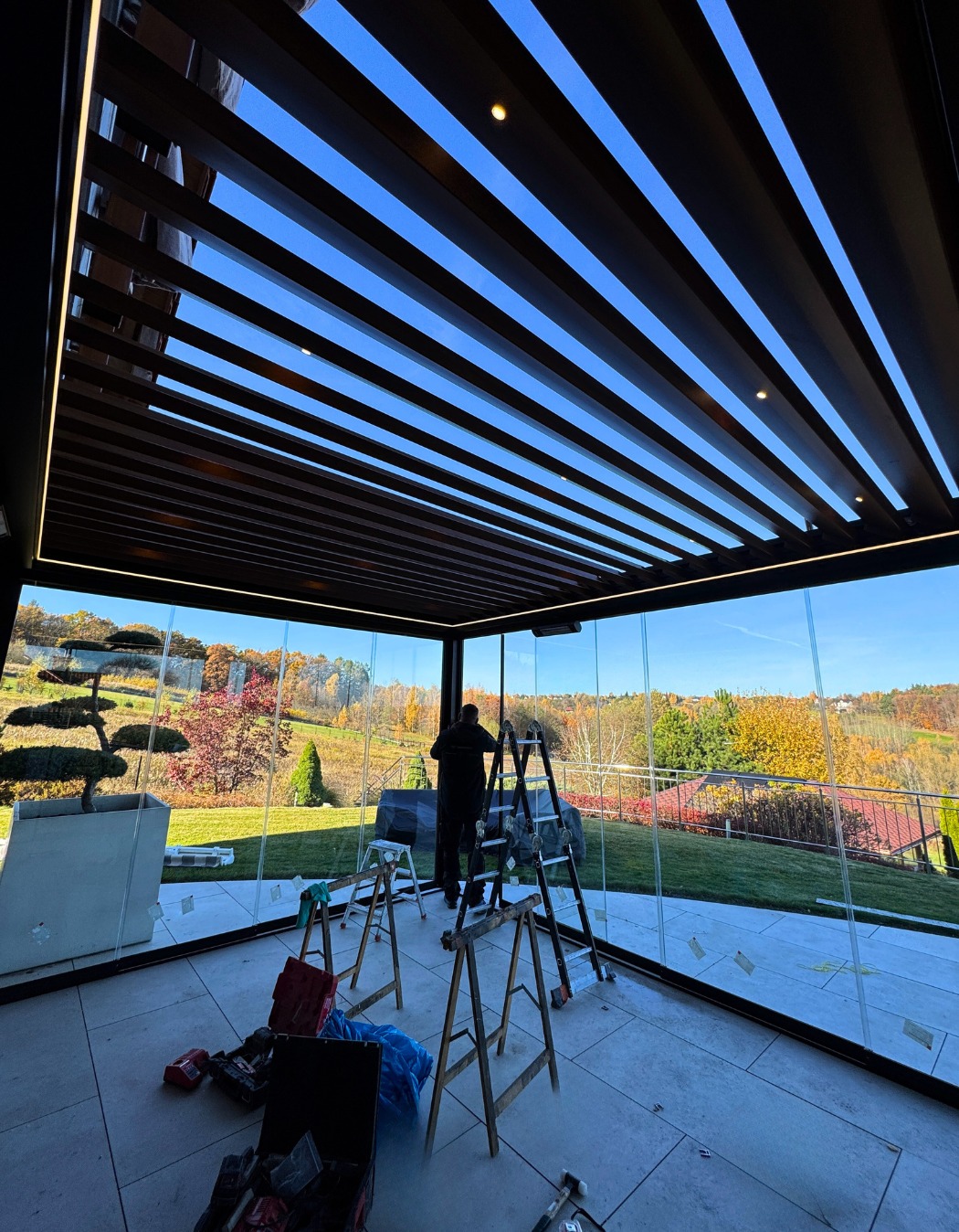 Nowoczesna zabudowa balkonu z przesuwnymi szklanymi panelami i drewnianą pergolą z oświetleniem LED. Widok na jesienny ogród. Prace montażowe w toku.