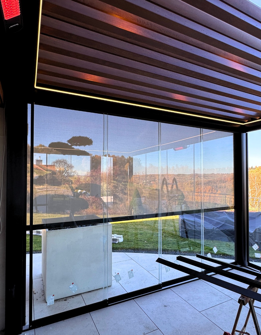 Nowoczesna zabudowa tarasu z przesuwnymi panelami szklanymi i roletami screen, widok na ogród. Oświetlenie LED podkreśla drewnianą konstrukcję pergoli.