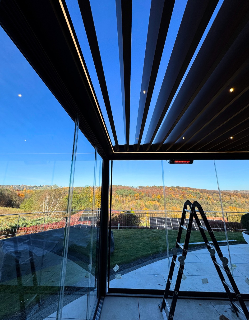 Nowoczesna zabudowa tarasu z czarną pergolą lamelową i szklanymi ścianami, widok na jesienny krajobraz. Drabina oparta o ścianę.