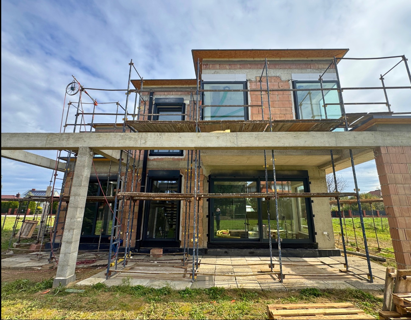 Montaż okien w surowym budynku. Widoczne rusztowania i niezakończone prace murarskie. Okna z czarnymi ramami, duże przeszklenia. Etap budowy.