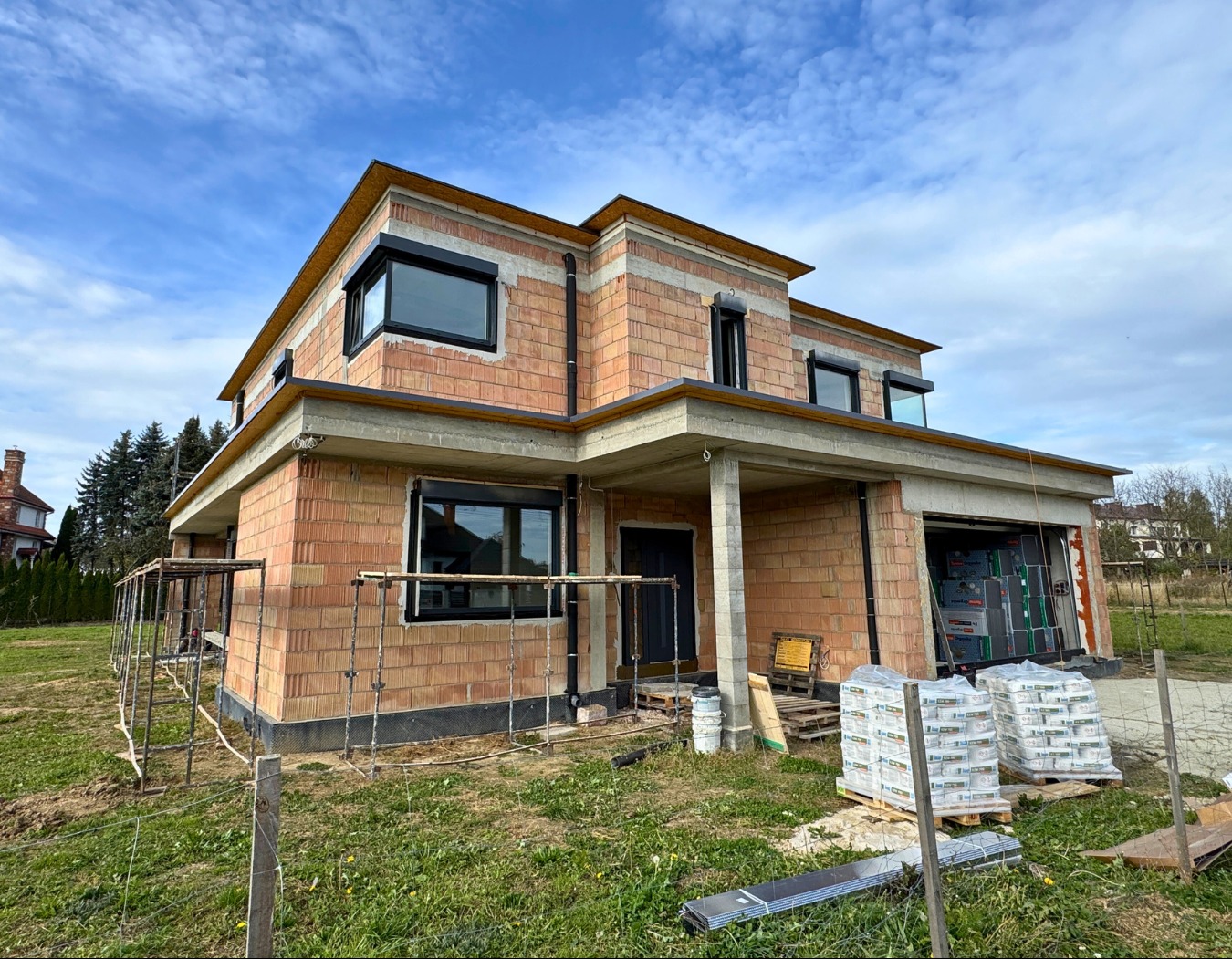 Surowy stan dwukondygnacyjnego domu z cegły z czarnymi oknami, widoczne materiały budowlane i rusztowania. Dom w trakcie budowy, z otwartym garażem.