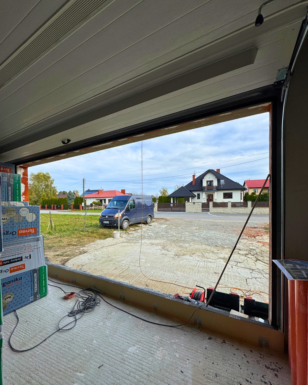 Montaż bramy garażowej segmentowej: widok z wnętrza garażu na podjazd i dom. Widoczne narzędzia i materiały budowlane. Brama w trakcie instalacji.