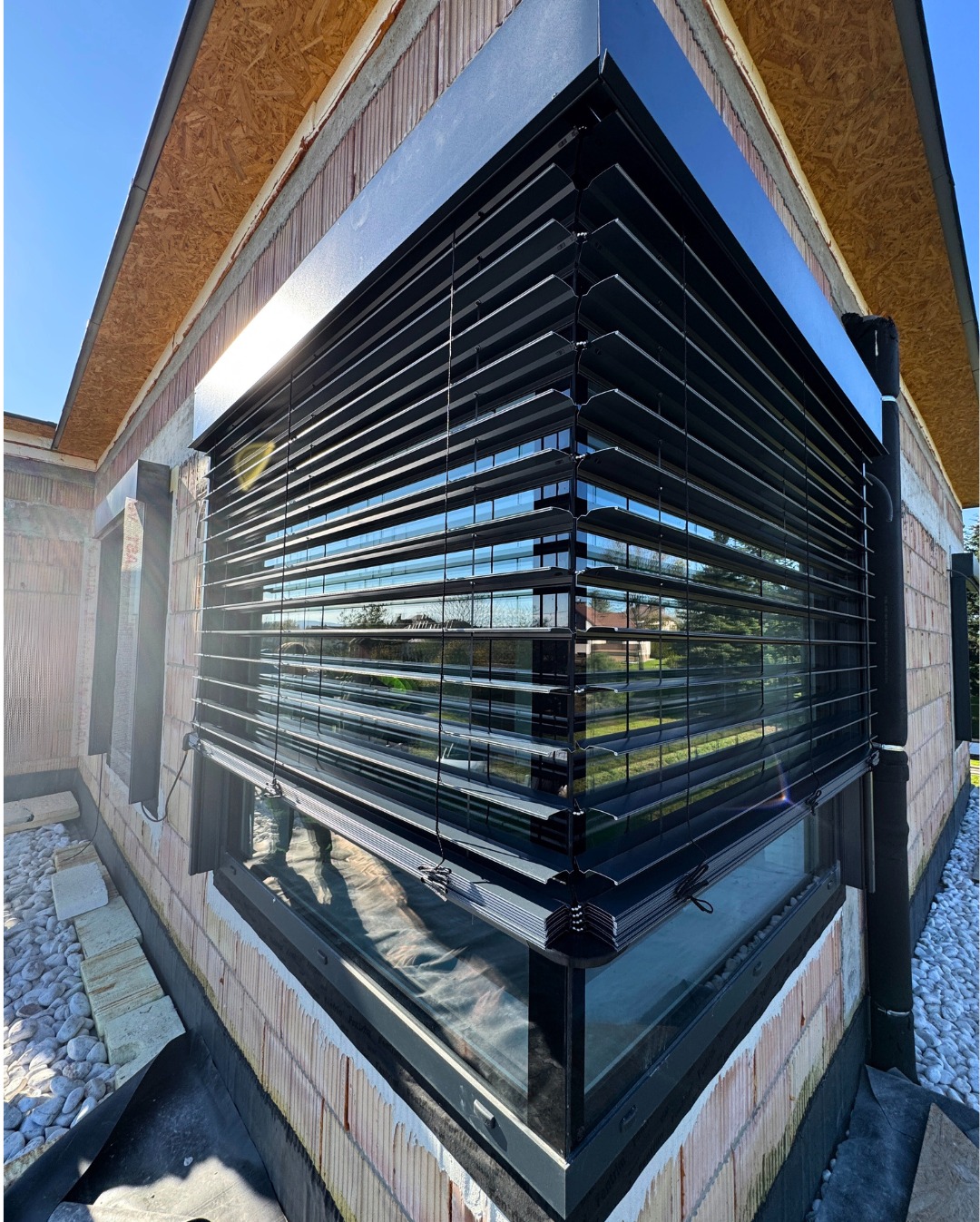 Nowoczesne, czarne żaluzje fasadowe zamontowane na oknie budynku w trakcie budowy, widoczne cegły i elementy konstrukcyjne. Częściowo opuszczone lamele.