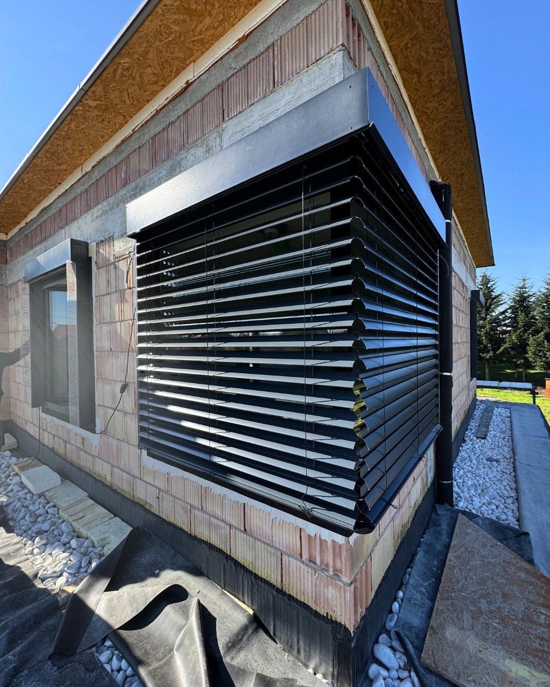 Zamontowane czarne żaluzje fasadowe na oknie budynku w trakcie budowy, widoczne białe kamienie i folia budowlana. Ujęcie z dołu, błękitne niebo.