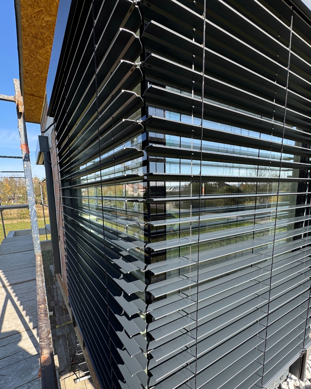 Elewacja budynku z zamontowanymi szarymi żaluzjami fasadowymi, widok z rusztowania. Widoczne odbicia w szybach i elementy konstrukcyjne.