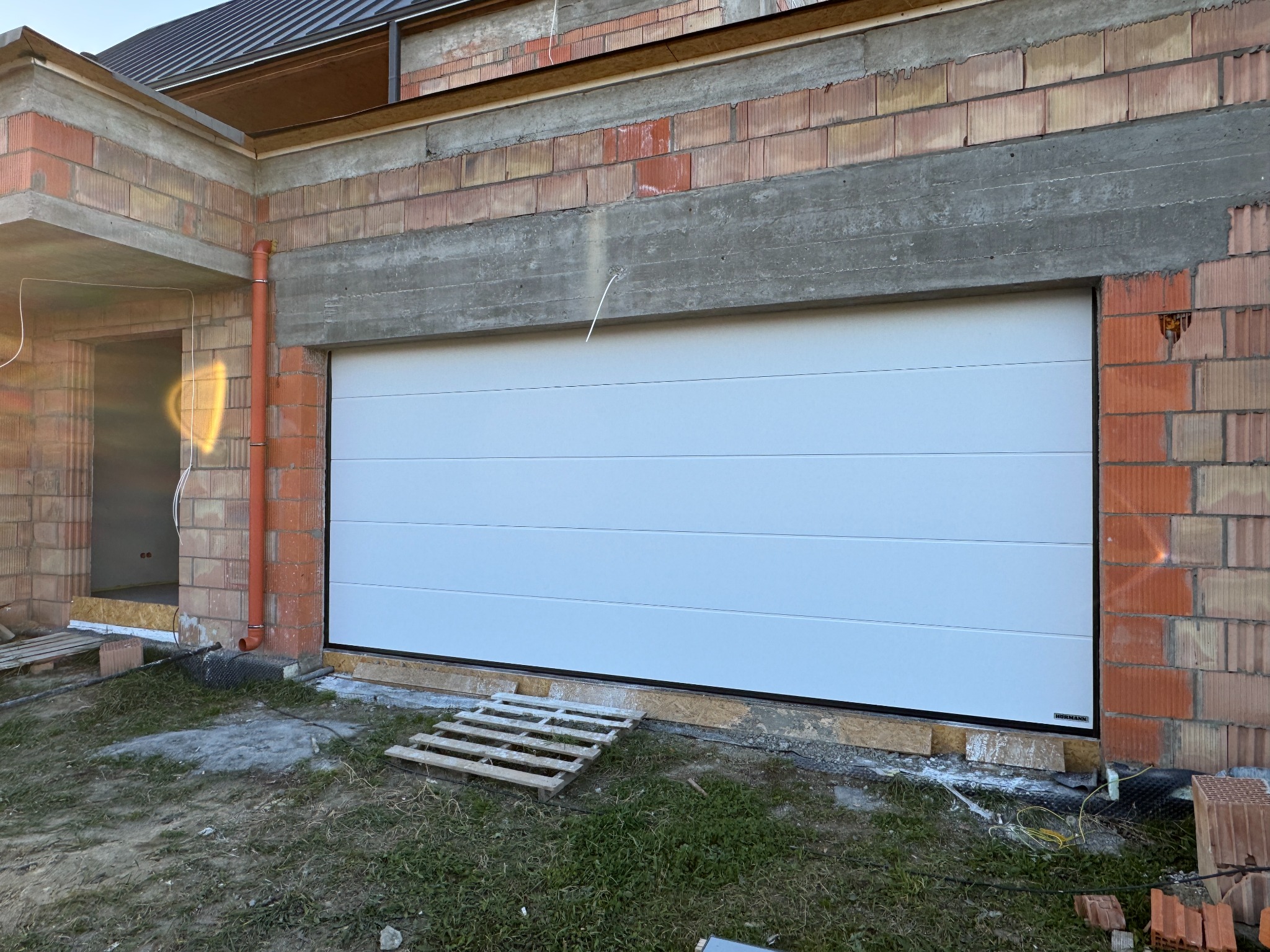 Biała segmentowa brama garażowa Hormann w surowym otoczeniu budowy, z widocznymi cegłami i elementami konstrukcyjnymi. Na dole leży drewniana paleta.
