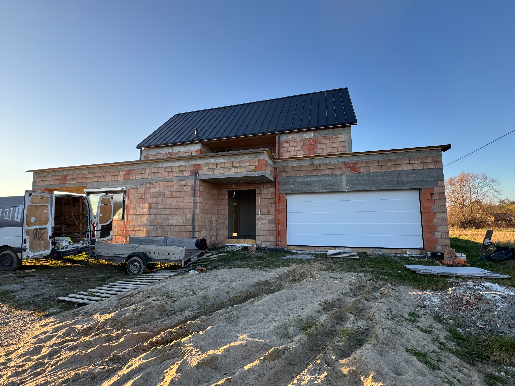 Nowoczesny dom w trakcie budowy z białą bramą garażową, widoczny z zewnątrz, z ceglanymi ścianami i czarnym dachem, na tle błękitnego nieba. Na podwórku stoi bus i przyczepka.