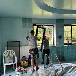 Polska Grupa Termomodernizacyjna - Montaż okna w pomieszczeniu z basenem. Dwóch mężczyzn w szarych koszulkach instaluje czarną ramę okienną. Widoczne narzędzia i akcesoria montażowe.