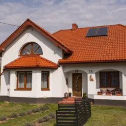 Biały dom jednorodzinny z czerwoną dachówką i panelami solarnymi na dachu, widok z przodu, zieleń w tle. Dom z poddaszem, drewniane okna i drzwi.