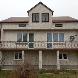 Polska Grupa Termomodernizacyjna - Dwupiętrowy dom z elewacją typu baranek, brązowym dachem i balkonem z metalową balustradą. Widoczna antena satelitarna i numer domu. Pochmurne niebo w tle.