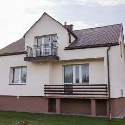 Polska Grupa Termomodernizacyjna - Dwukondygnacyjny dom z balkonem i brązowym dachem, numer posesji '50' widoczny na elewacji. Detale: antena na balkonie, brązowy cokół budynku, zielony trawnik.