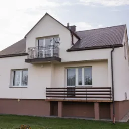 Dwukondygnacyjny dom z balkonem i brązowym dachem, numer posesji '50' widoczny na elewacji. Detale: antena na balkonie, brązowy cokół budynku, zielony trawnik.