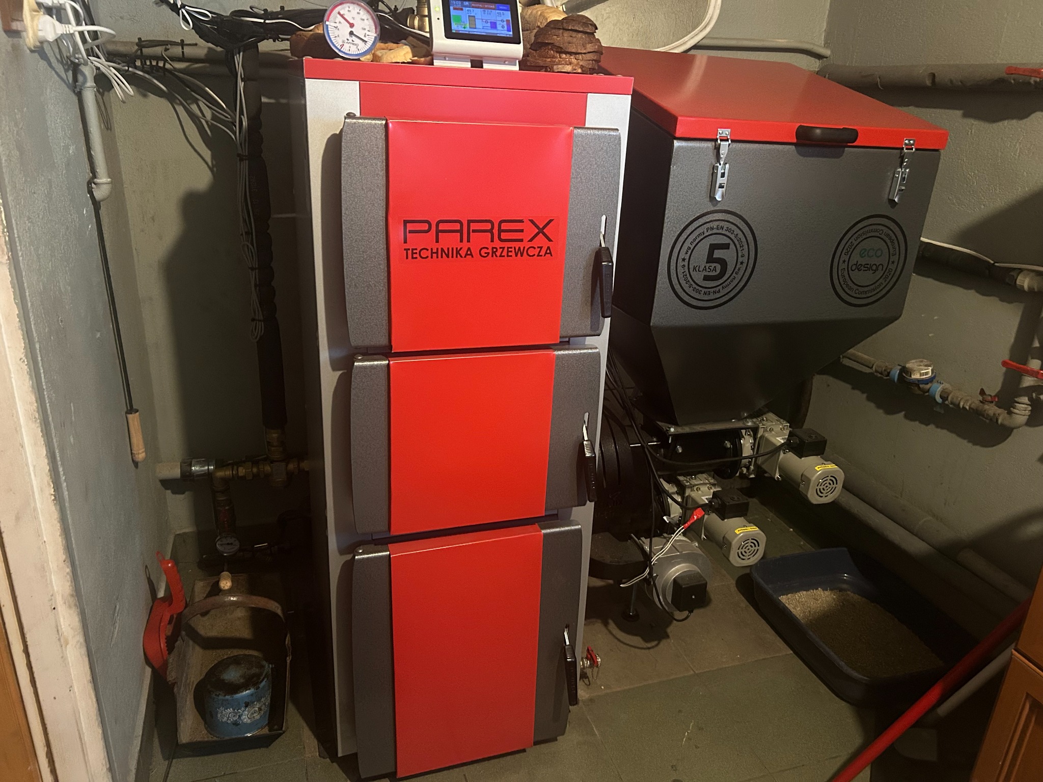 Nowoczesny piec na paliwo stałe marki Parex z podajnikiem, oznaczony klasą 5 i etykietą Eco Design, zainstalowany w kotłowni. Widoczne przewody i elementy instalacji.