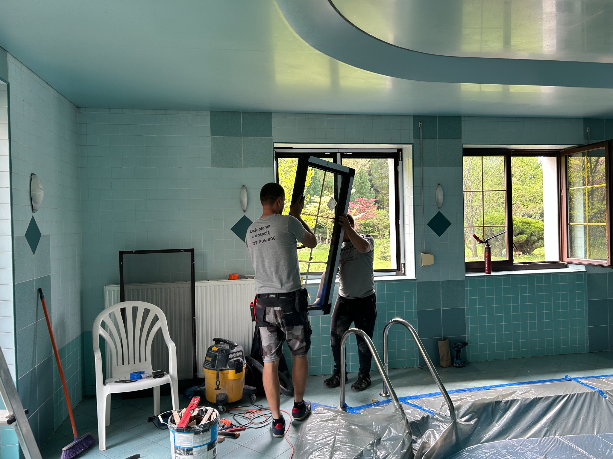 Montaż okna w pomieszczeniu z basenem. Dwóch mężczyzn w szarych koszulkach instaluje czarną ramę okienną. Widoczne narzędzia i akcesoria montażowe.