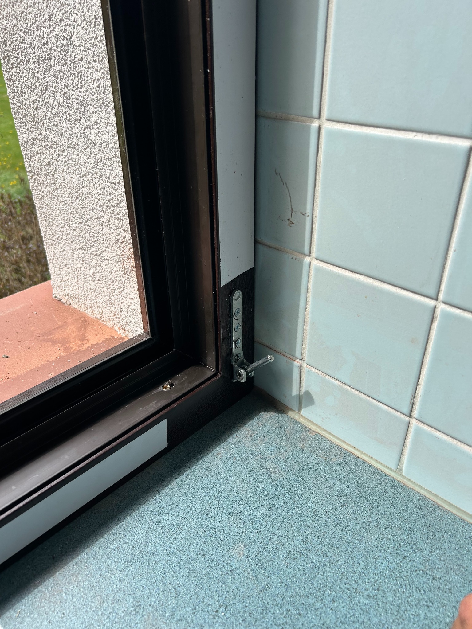 Szczegółowe ujęcie okna PCV z brązową ramą i zamontowanym okuciem w rogu, na tle niebieskich płytek i ściany z widoczną fakturą.