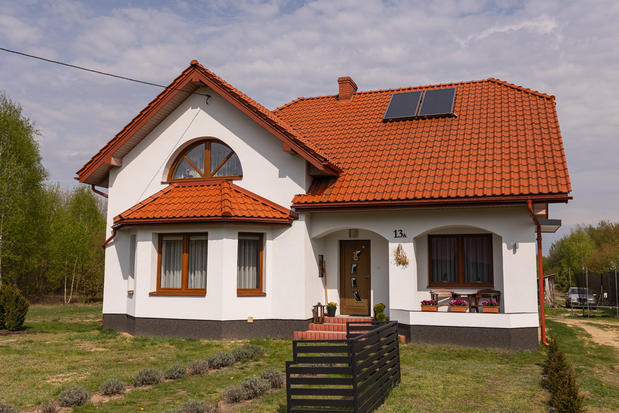 Biały dom jednorodzinny z czerwoną dachówką i panelami solarnymi na dachu, widok z przodu, zieleń w tle. Dom z poddaszem, drewniane okna i drzwi.