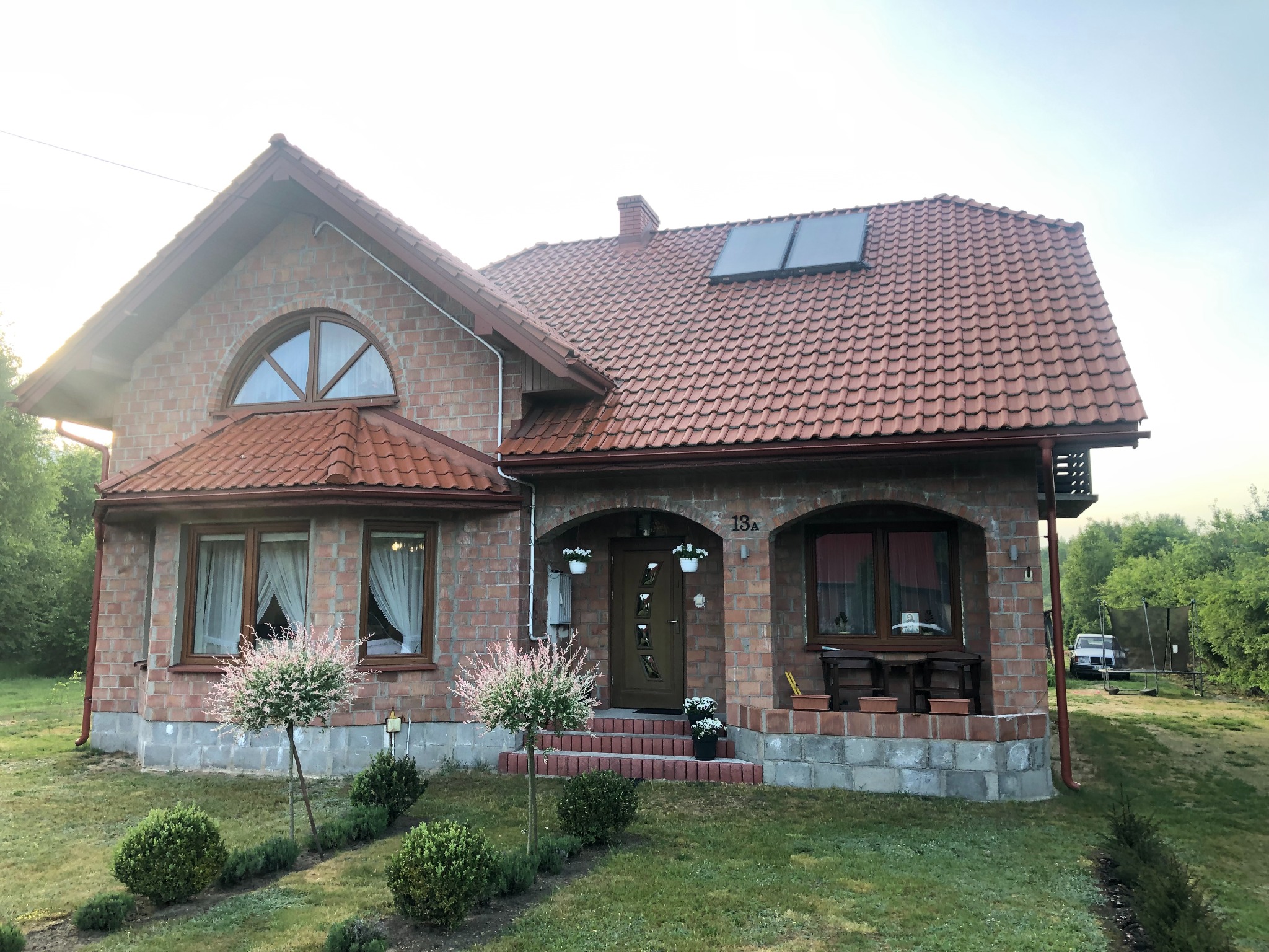 Dwupiętrowy dom z cegły klinkierowej z czerwonym dachem i panelami słonecznymi. Widoczny ogród z roślinami i drzewami. Dom z podjazdem i numerem 13A nad wejściem.