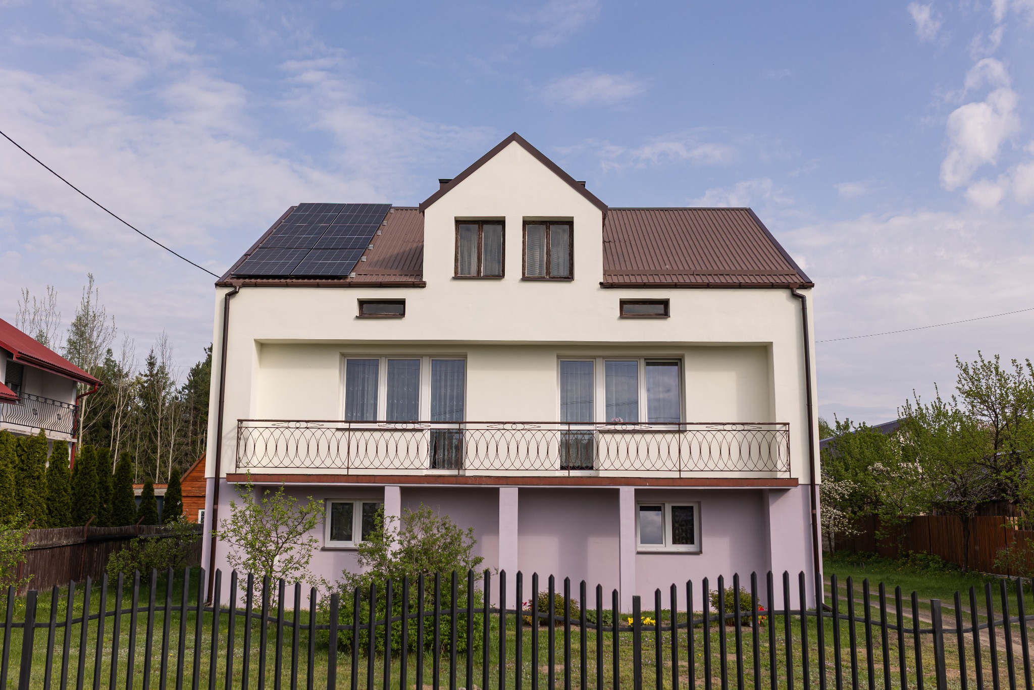 Dwupiętrowy dom z balkonem i panelami słonecznymi na brązowym dachu, otoczony zielenią i czarnym ogrodzeniem. Jasna elewacja z elementami w odcieniach fioletu.