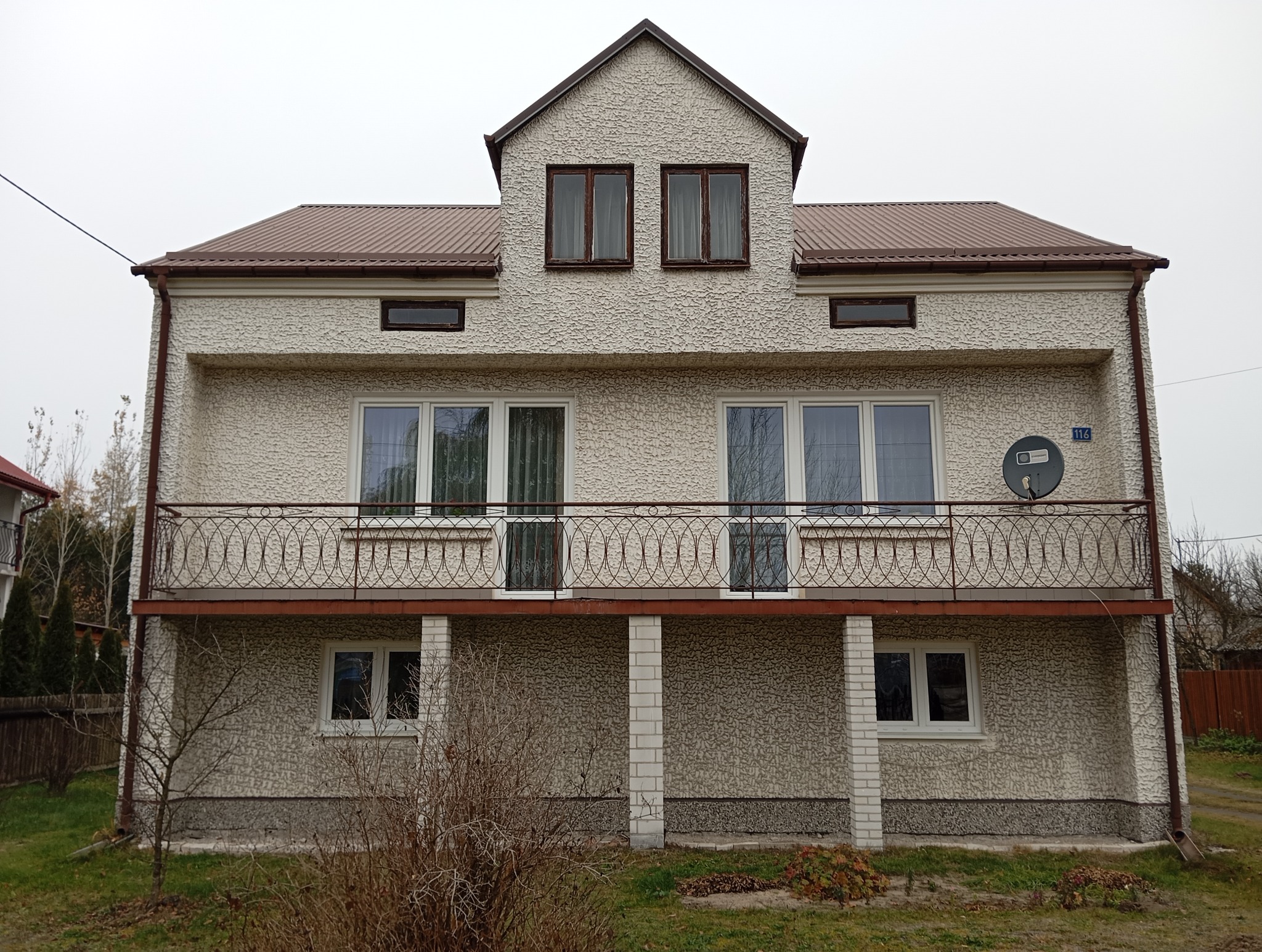 Dwupiętrowy dom z elewacją typu baranek, brązowym dachem i balkonem z metalową balustradą. Widoczna antena satelitarna i numer domu. Pochmurne niebo w tle.