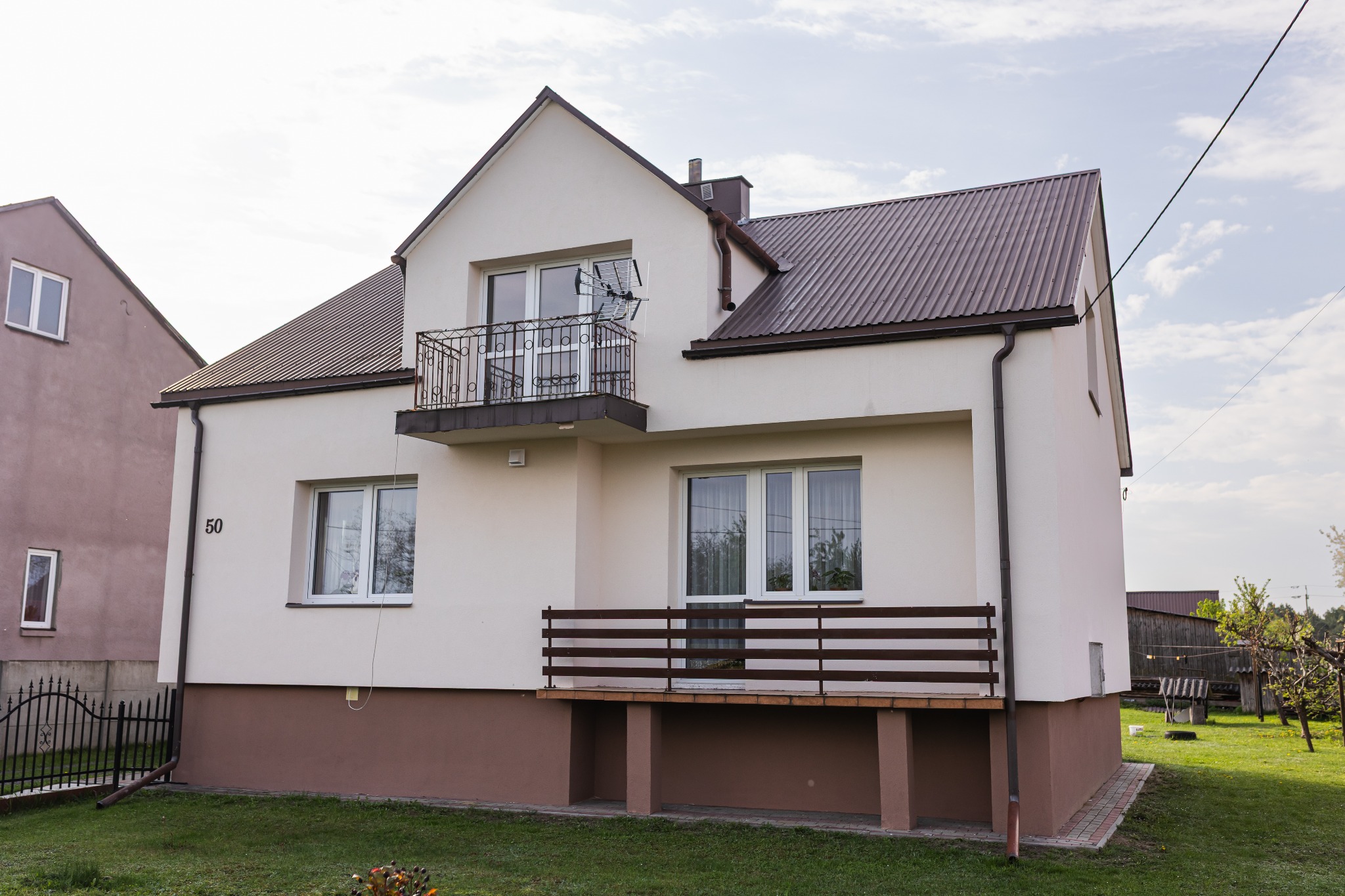 Dwukondygnacyjny dom z balkonem i brązowym dachem, numer posesji '50' widoczny na elewacji. Detale: antena na balkonie, brązowy cokół budynku, zielony trawnik.