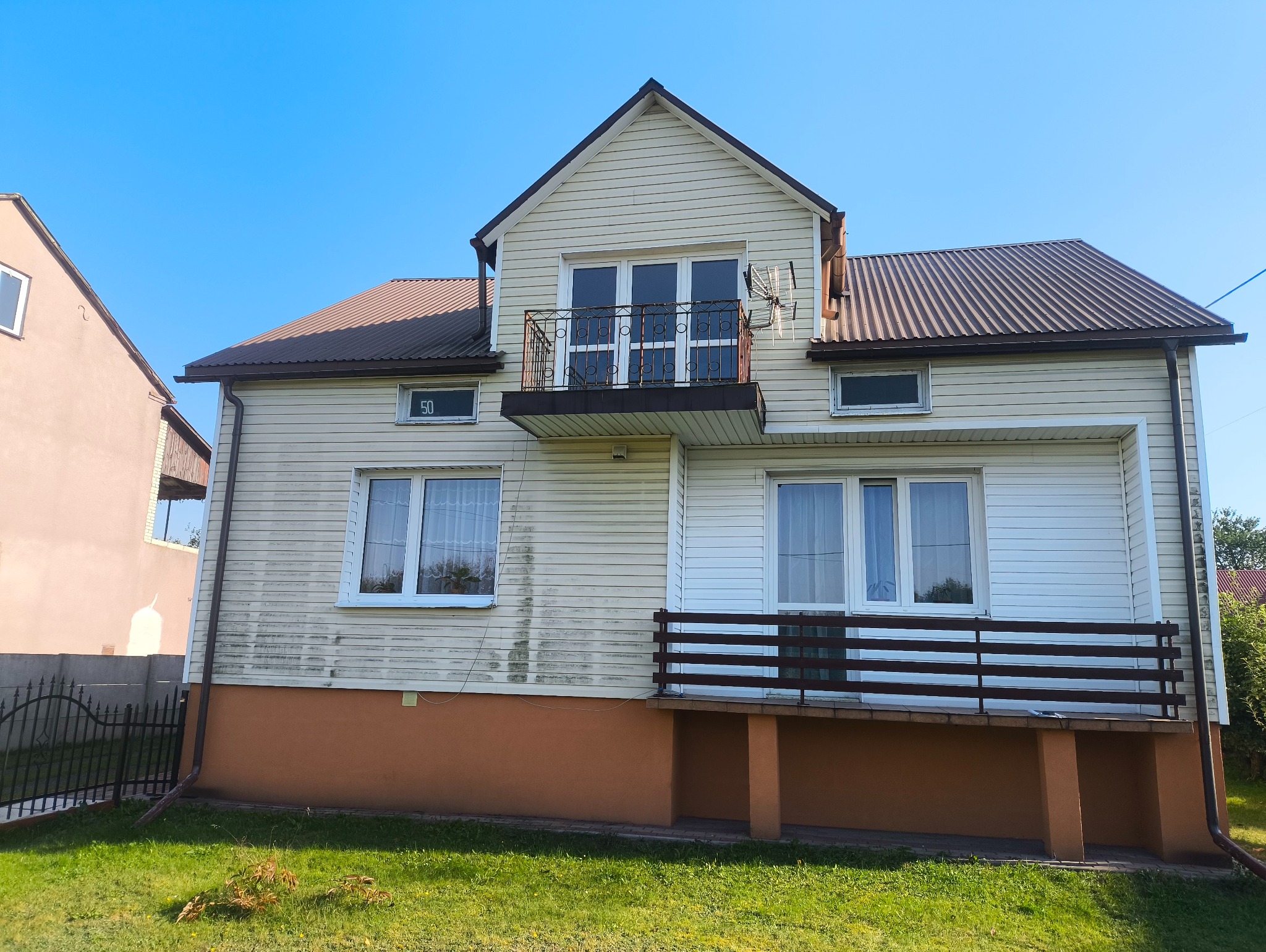 Dom z sidingiem i balkonem, widoczne ślady zabrudzeń na elewacji, brązowy dach i podmurówka. Na ścianie numer domu '50'.