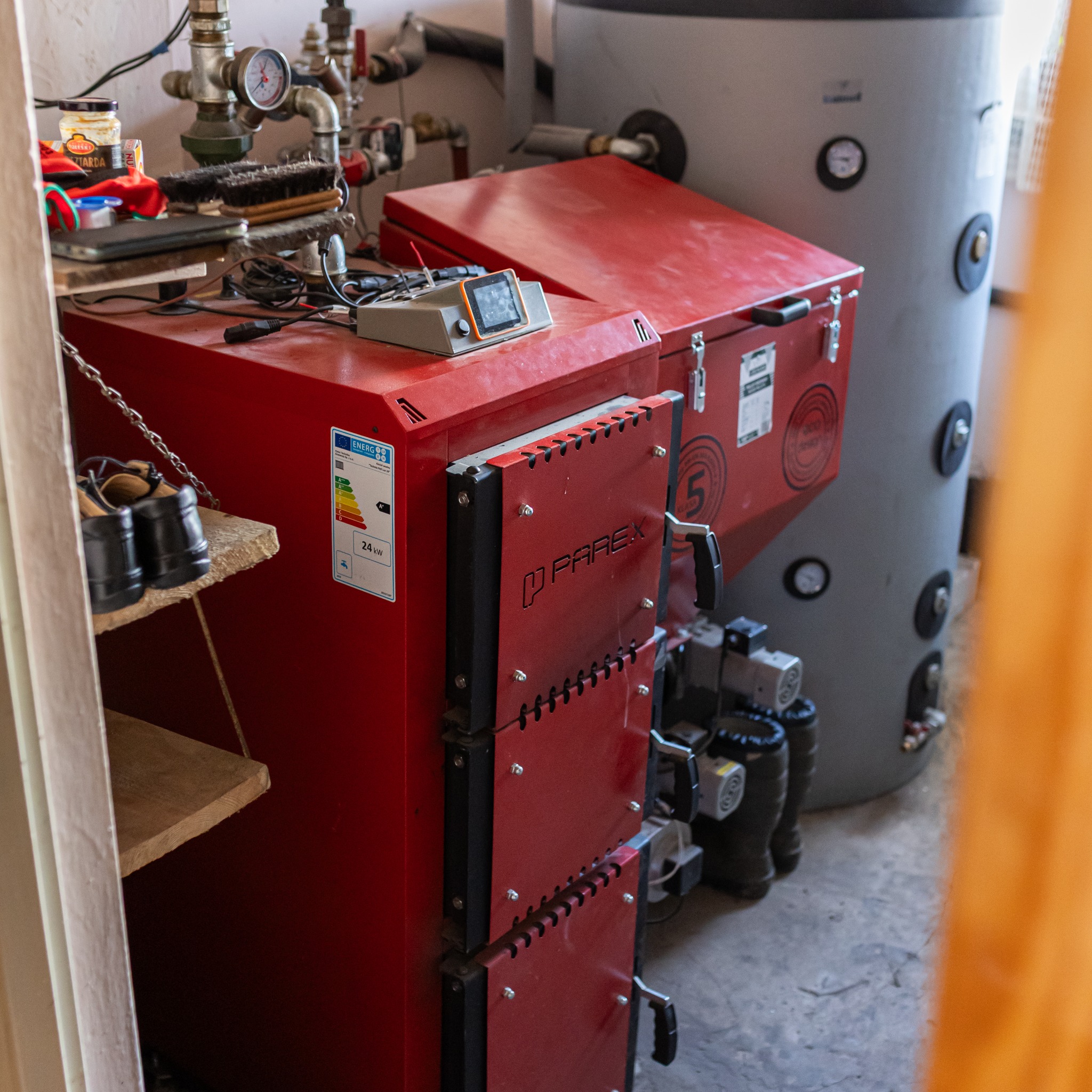 Czerwony piec marki Parex z etykietą energetyczną 24kW, obok szary zbiornik akumulacyjny. Na piecu leży sterownik. W tle widoczne instalacje i akcesoria.
