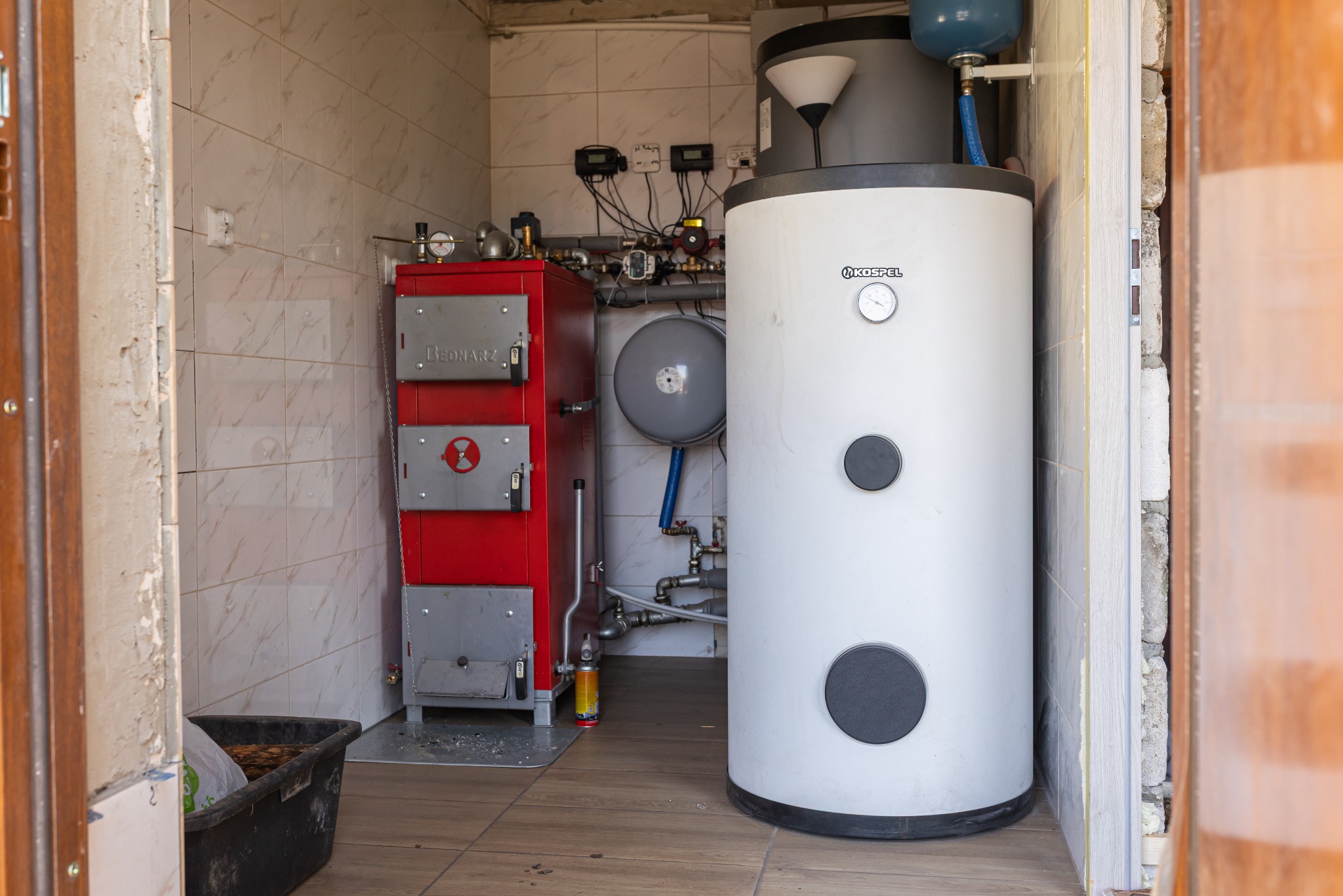 Kotłownia z piecem na paliwo stałe 'Bednarz' i bojlerem Kospel, widoczne rury instalacji CO, całość w pomieszczeniu wyłożonym płytkami ceramicznymi.
