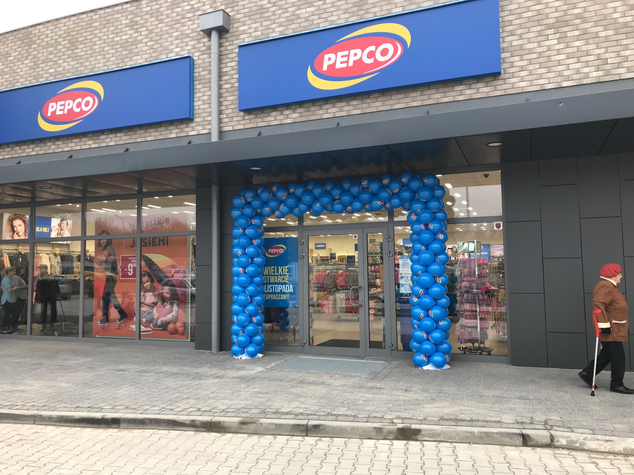 Wejście do sklepu Pepco ozdobione łukiem z niebieskich balonów, szyldy z logo Pepco nad wejściem, osoba z laską przechodząca obok.