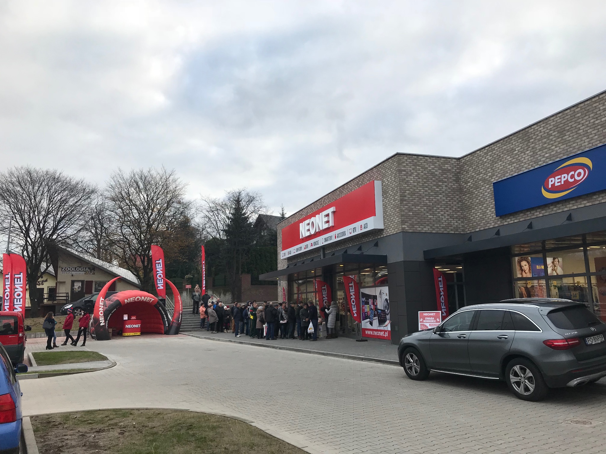Długa kolejka klientów przed sklepem Neonet i Pepco w pochmurny dzień, z reklamowymi bramami i flagami Neonet ustawionymi na zewnątrz.