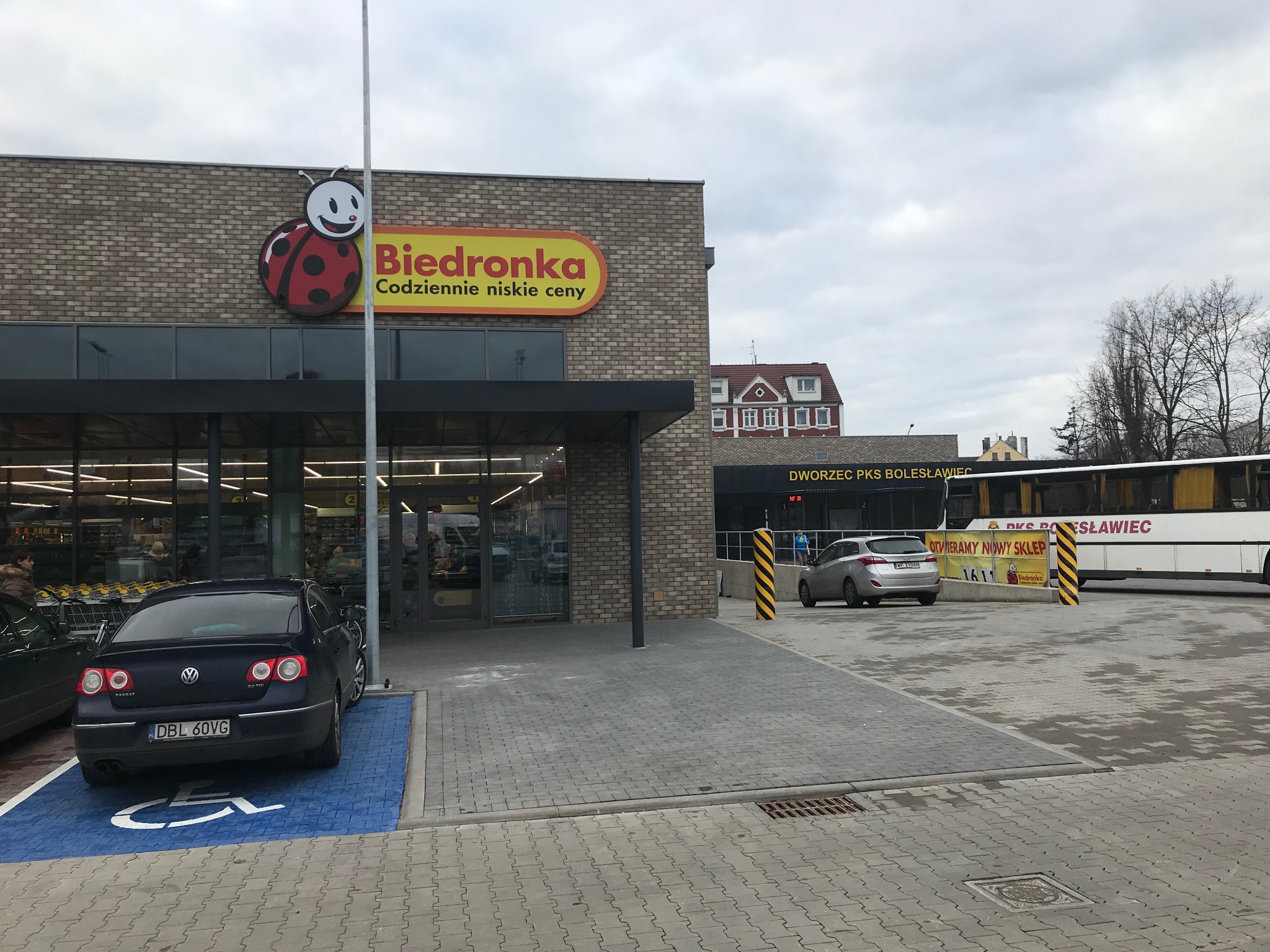 Budynek sklepu Biedronka z ceglaną elewacją, widoczny znak firmowy z uśmiechniętą biedronką, zaparkowany samochód na miejscu dla niepełnosprawnych i autobus PKS w tle.
