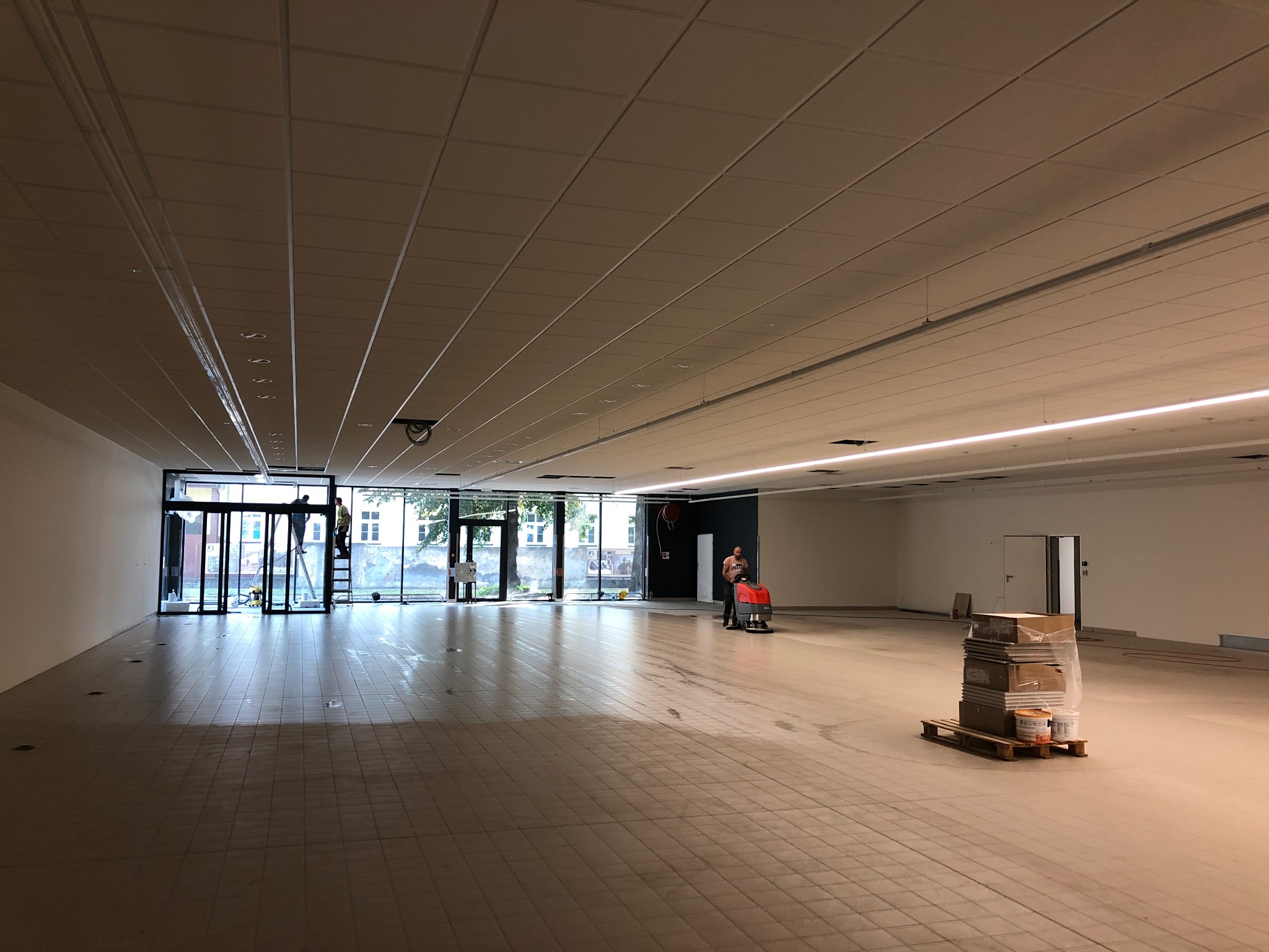 Wnętrze hali w trakcie prac wykończeniowych: pracownik myje posadzkę maszyną czyszczącą, paleta z materiałami budowlanymi w rogu, widoczne okna i drabina z pracownikami na zewnątrz.