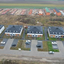 TRIDENT HOUSE - Sp. z o.o. - Widok z góry na osiedle nowych domów jednorodzinnych z czarnymi dachami i białymi elewacjami, z zaparkowanymi samochodami i placem budowy w tle, gdzie widać fundamenty kolejnych budynków.