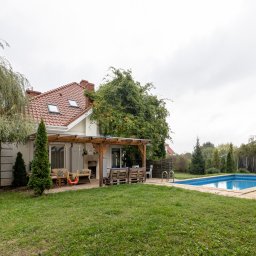 TRIDENT HOUSE - Sp. z o.o. - Dom jednorodzinny z ogrodem, basenem i zadaszonym tarasem z meblami ogrodowymi i huśtawką dla dzieci, otoczony zielenią i drzewami.