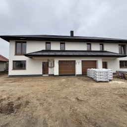 TRIDENT HOUSE - Sp. z o.o. - Bliźniak w stanie deweloperskim z brązowymi bramami garażowymi i oknami, na działce leżą palety z materiałami budowlanymi.