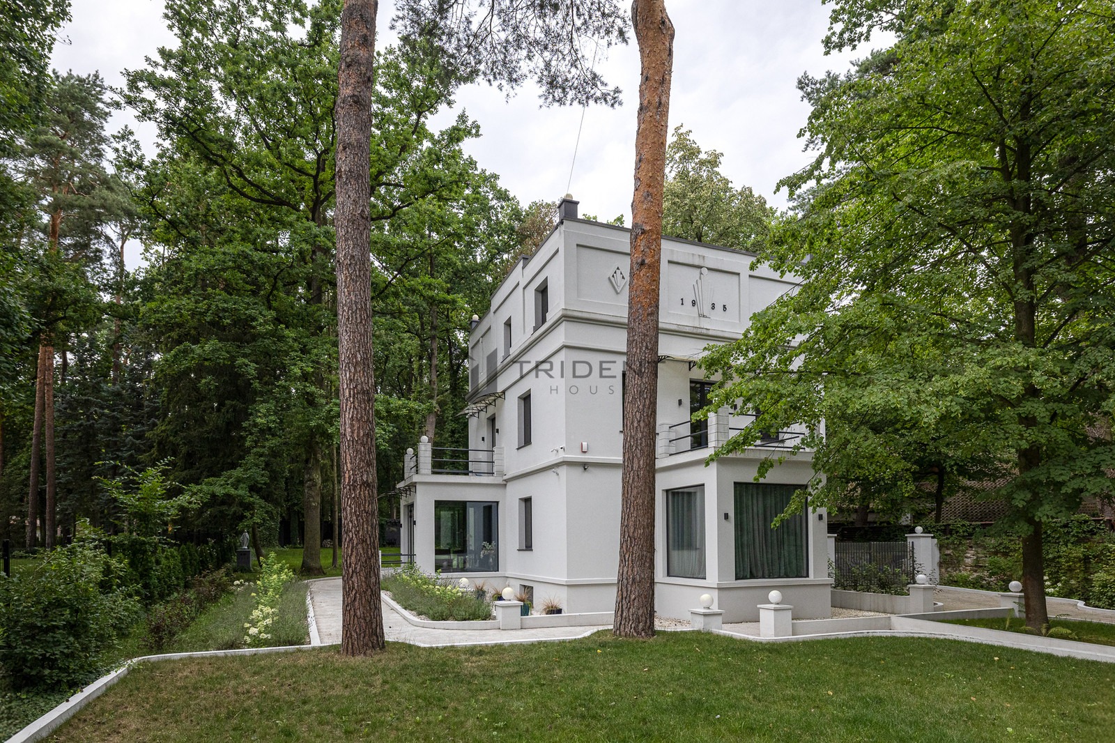 Trzypiętrowy, biały dom z balkonami, otoczony drzewami i trawnikiem, z napisem 'TRIDE HOUSE' i rokiem '1935' na fasadzie.