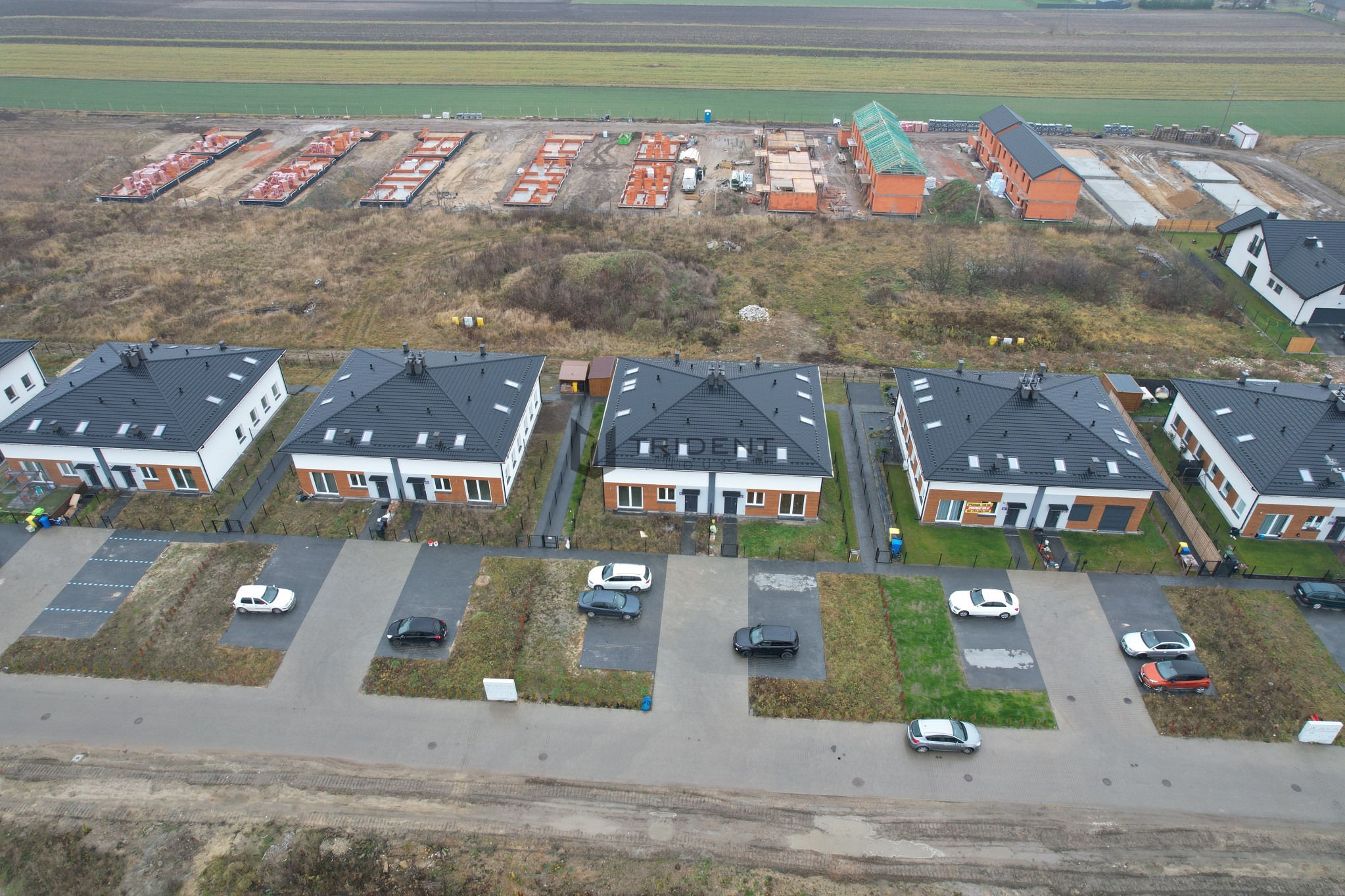 Widok z góry na osiedle nowych domów jednorodzinnych z czarnymi dachami i białymi elewacjami, z zaparkowanymi samochodami i placem budowy w tle, gdzie widać fundamenty kolejnych budynków.