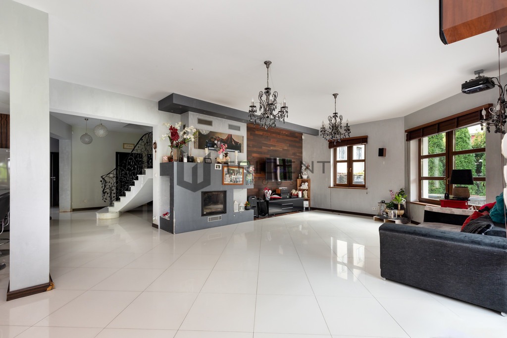 Przestronny salon z białymi płytkami podłogowymi, kominkiem obudowanym szarym kamieniem, drewnianą ścianą telewizyjną i schodami z ozdobną, kutą balustradą.