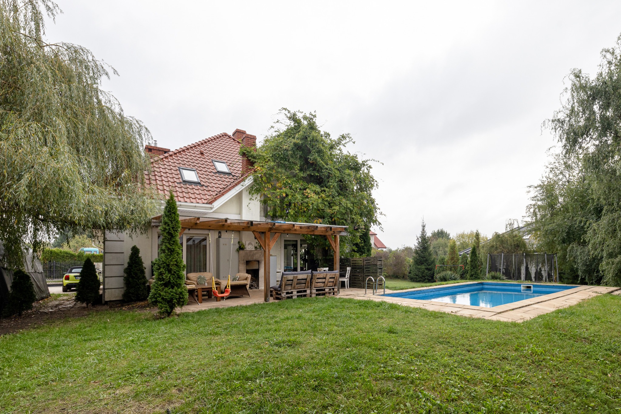 Dom jednorodzinny z ogrodem, basenem i zadaszonym tarasem z meblami ogrodowymi i huśtawką dla dzieci, otoczony zielenią i drzewami.