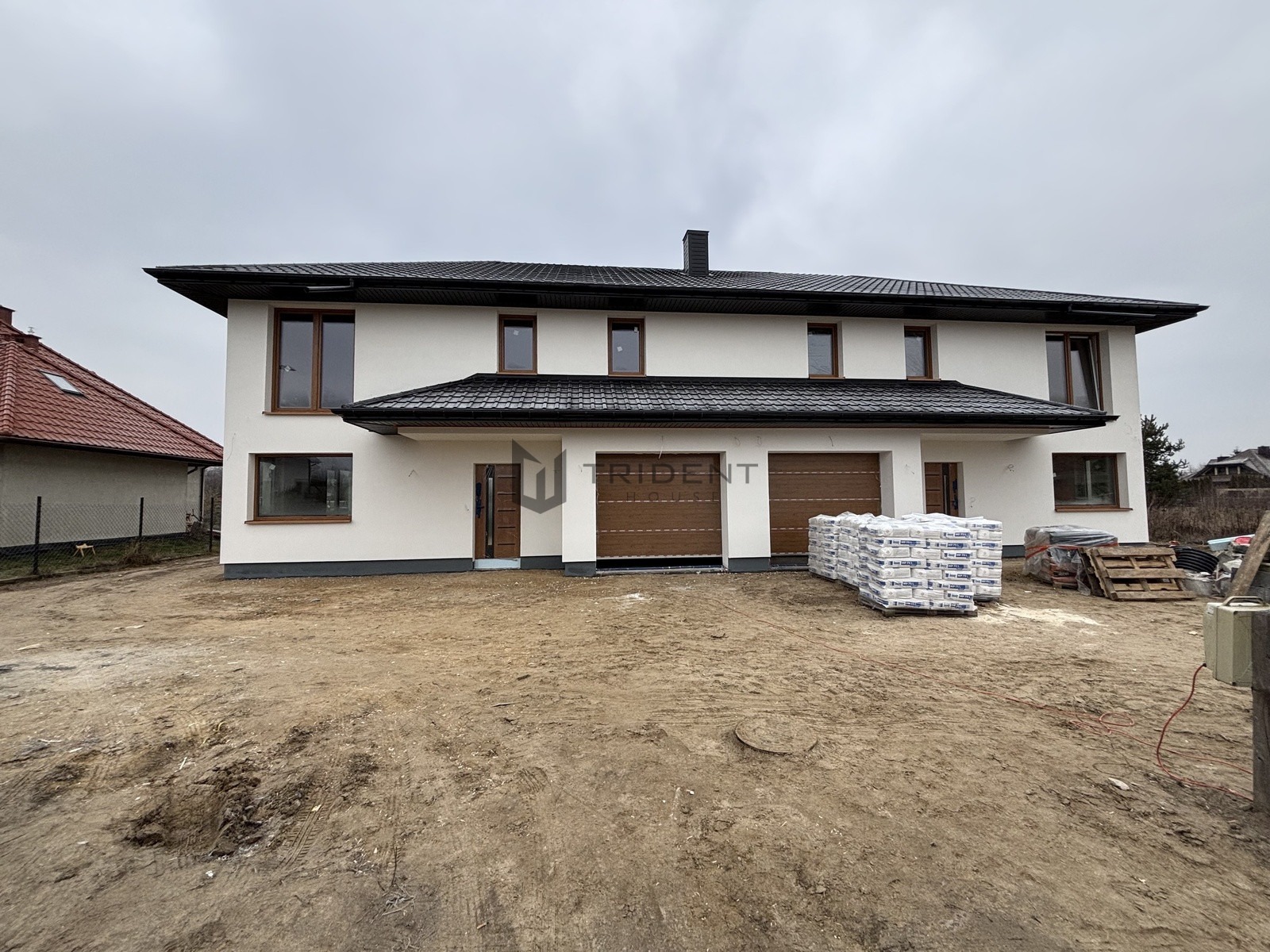 Bliźniak w stanie deweloperskim z brązowymi bramami garażowymi i oknami, na działce leżą palety z materiałami budowlanymi.