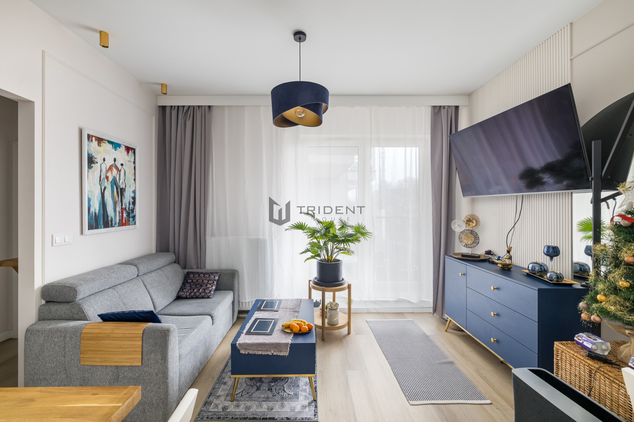 Nowoczesny salon z szarą sofą, niebieską komodą i stolikiem kawowym, dekoracjami i choinką w rogu, oświetlony lampą wiszącą z niebieskim abażurem.