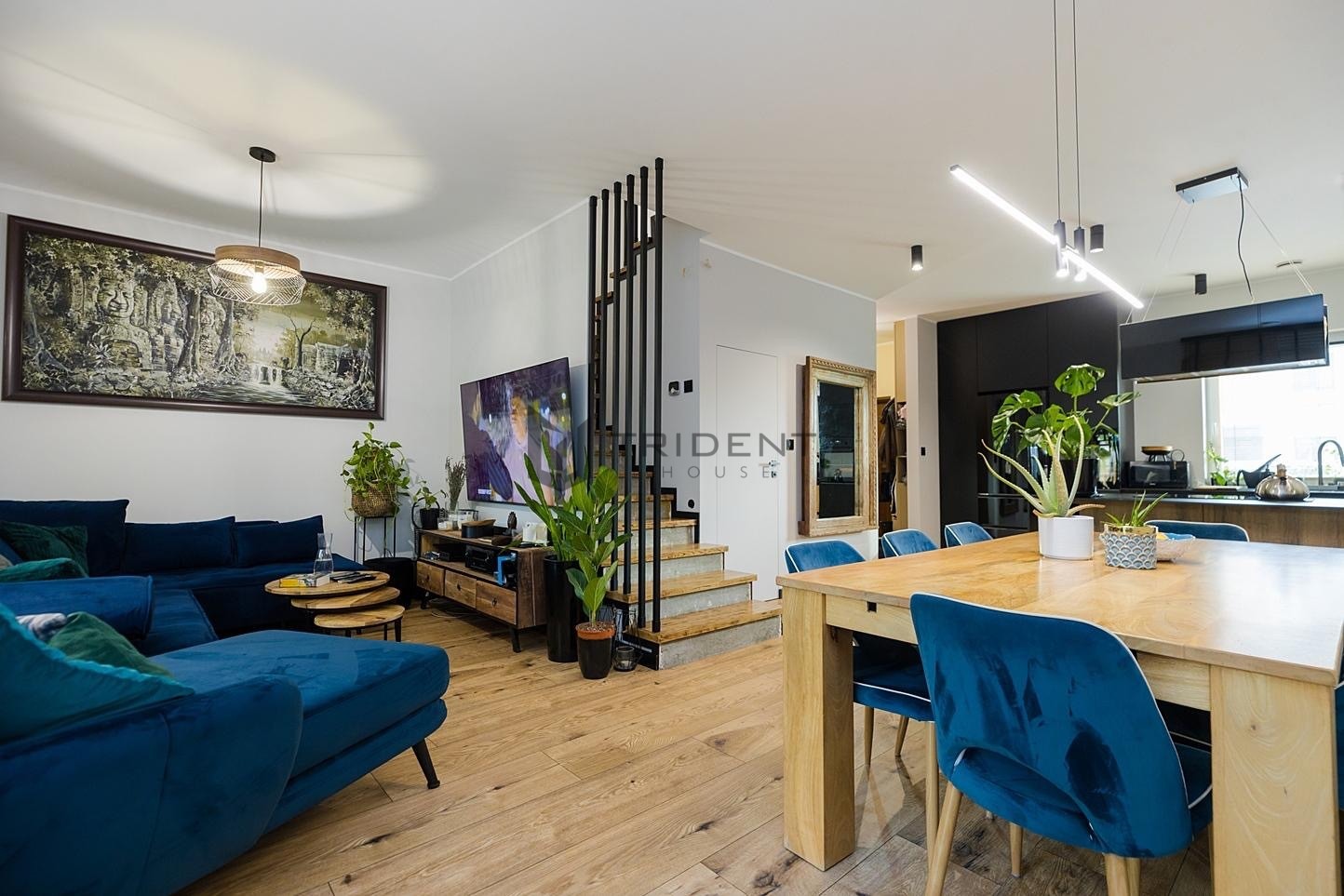 Przestronne wnętrze salonu z jadalnią w odcieniach błękitu i drewna, z dużym obrazem na ścianie i schodami z logo firmy.