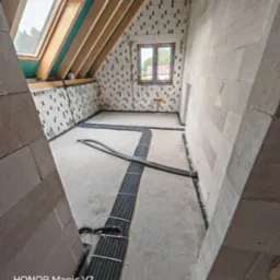 Instalacja przewodów w nowym budynku z oknem dachowym. Widoczne kable ułożone na podłodze, ściany z bloków. W tle okno z widokiem na drzewa.