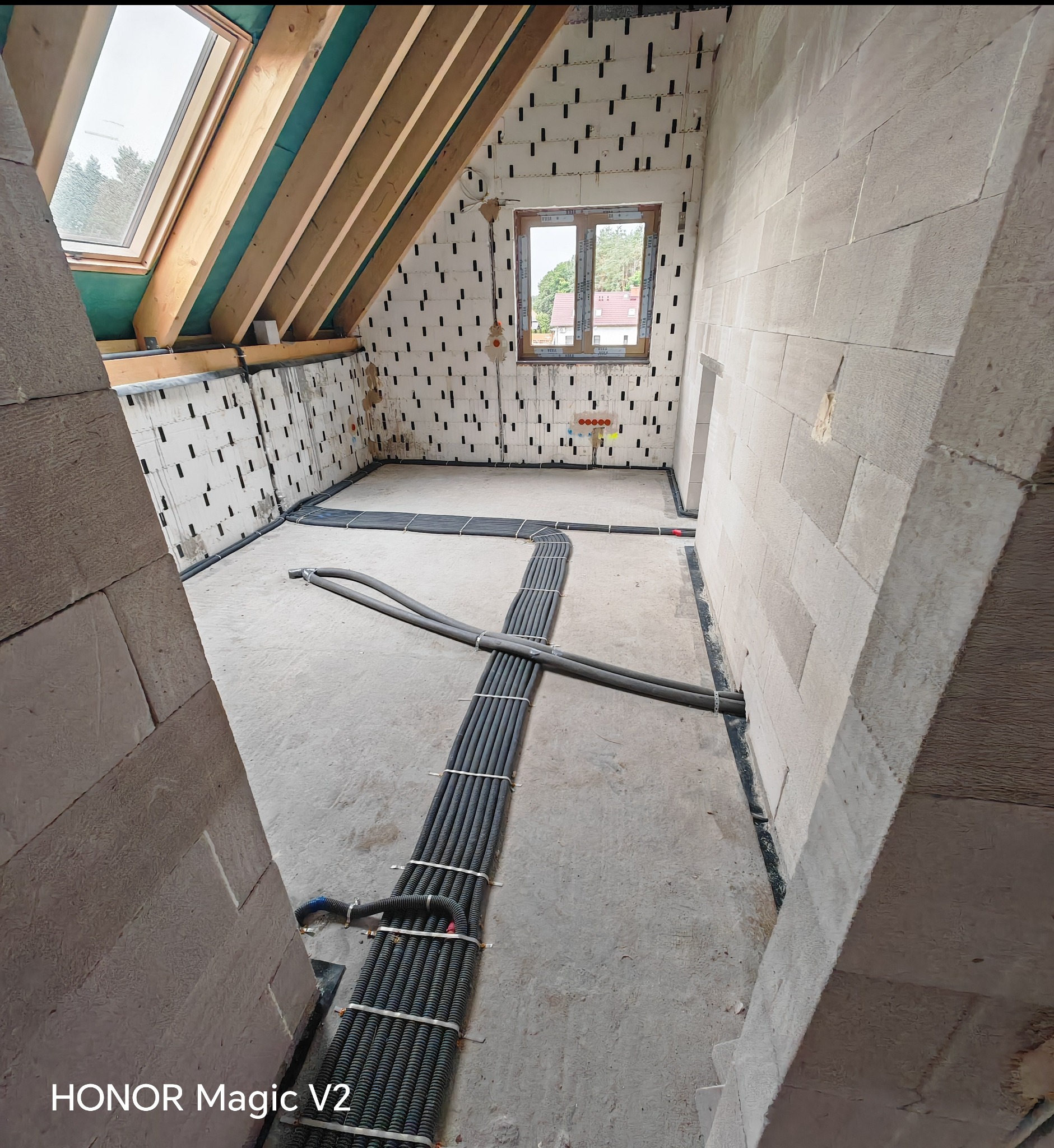 Instalacja przewodów w nowym budynku z oknem dachowym. Widoczne kable ułożone na podłodze, ściany z bloków. W tle okno z widokiem na drzewa.