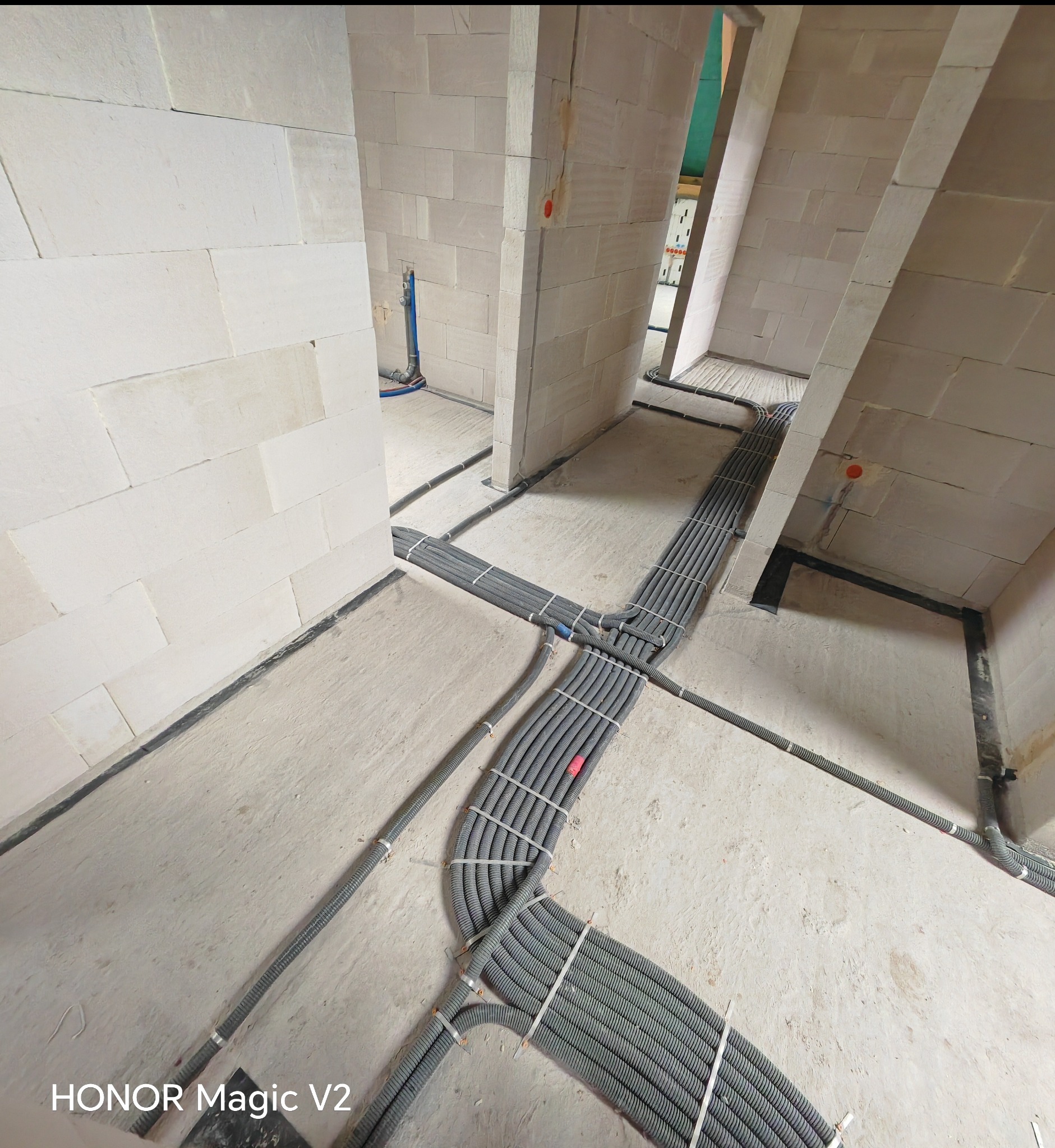 Rozprowadzenie przewodów w peszlach na podłodze w surowym budynku z białych bloczków, widoczne kable w ścianach. Instalacja elektryczna w trakcie realizacji.