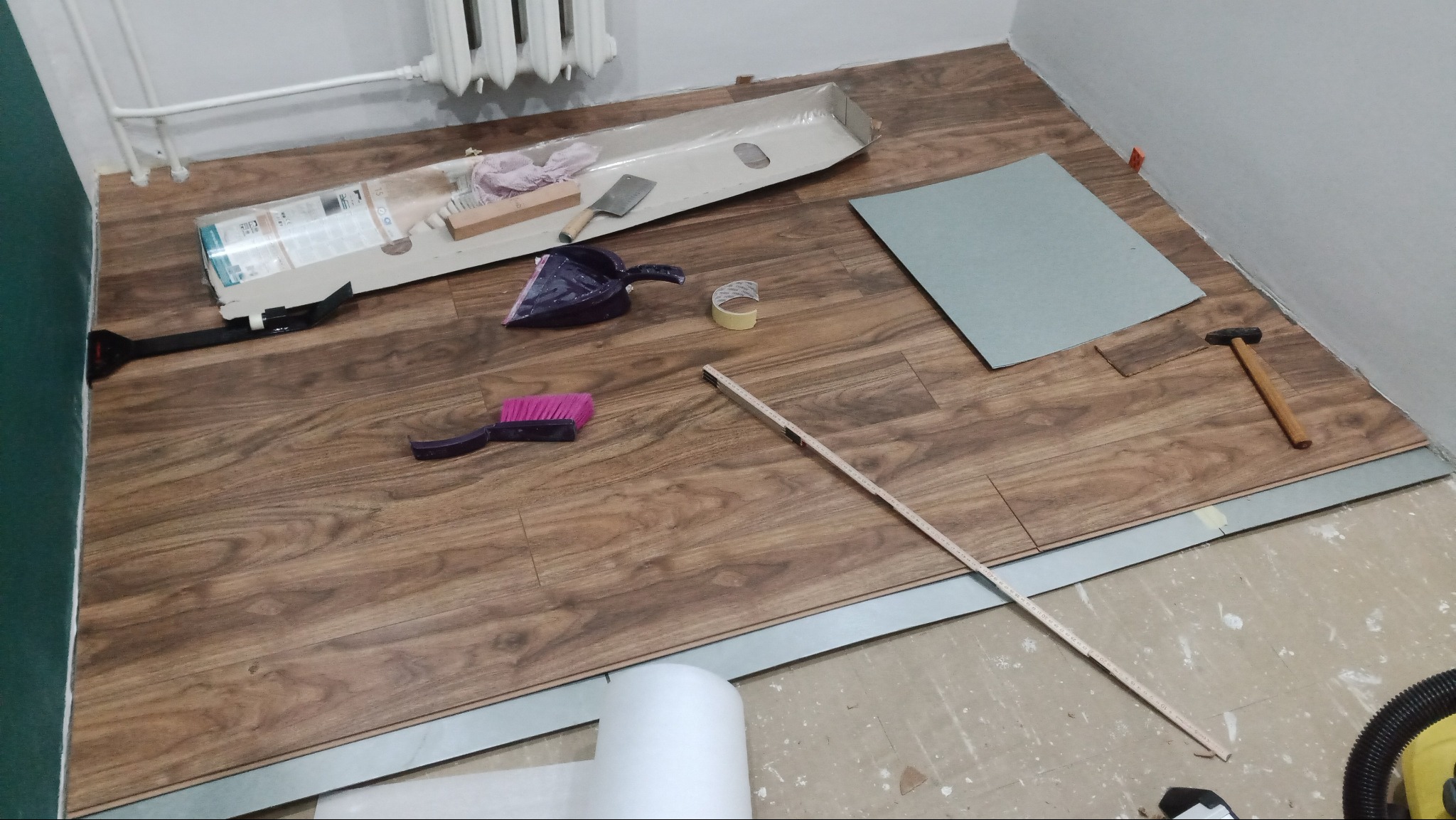 Fragment podłogi w trakcie układania paneli: widoczne panele z imitacją drewna, listwy przypodłogowe, narzędzia (młotek, miarka, szczotka, szpachelka), opakowanie paneli i podkład. Część podłogi...