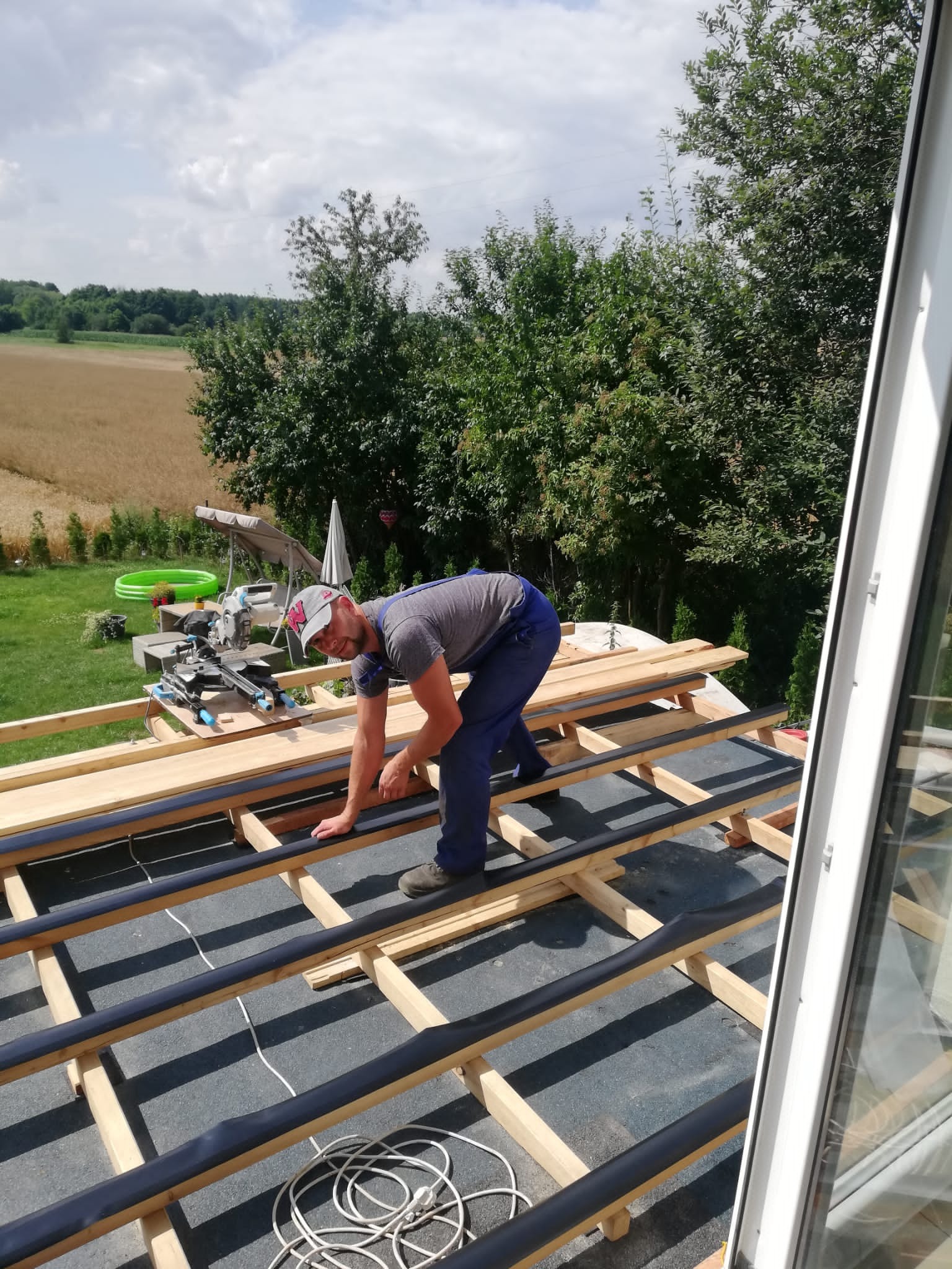 Uśmiechnięty mężczyzna w kombinezonie roboczym układa drewniane legary na dachu, widoczny fragment dachu z papą i izolacją, w tle zielony trawnik, drzewa i pole uprawne.