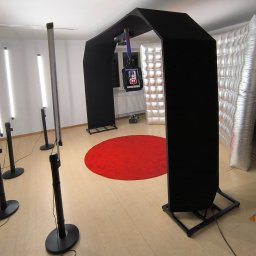 FOTOFRAJDA ALEKSANDRA MICEK - Wnętrze z rozstawioną czarną fotobudką z ekranem, oświetleniem LED i dmuchanym tłem, czerwony dywan na podłodze.