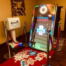 FOTOFRAJDA ALEKSANDRA MICEK - Nowoczesna fotobudka lustrzana z oświetleniem LED i napisem 'Lustro drukuje TWOJE ZDJĘCIE', otoczona słupkami z czerwonymi sznurami, obok biała drukarka do zdjęć i rekwizyty na czerwonym dywanie.