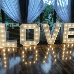 FOTOFRAJDA ALEKSANDRA MICEK - Świecący napis LOVE na parkiecie, duże litery z żarówkami, w tle okno z firankami i zielenią